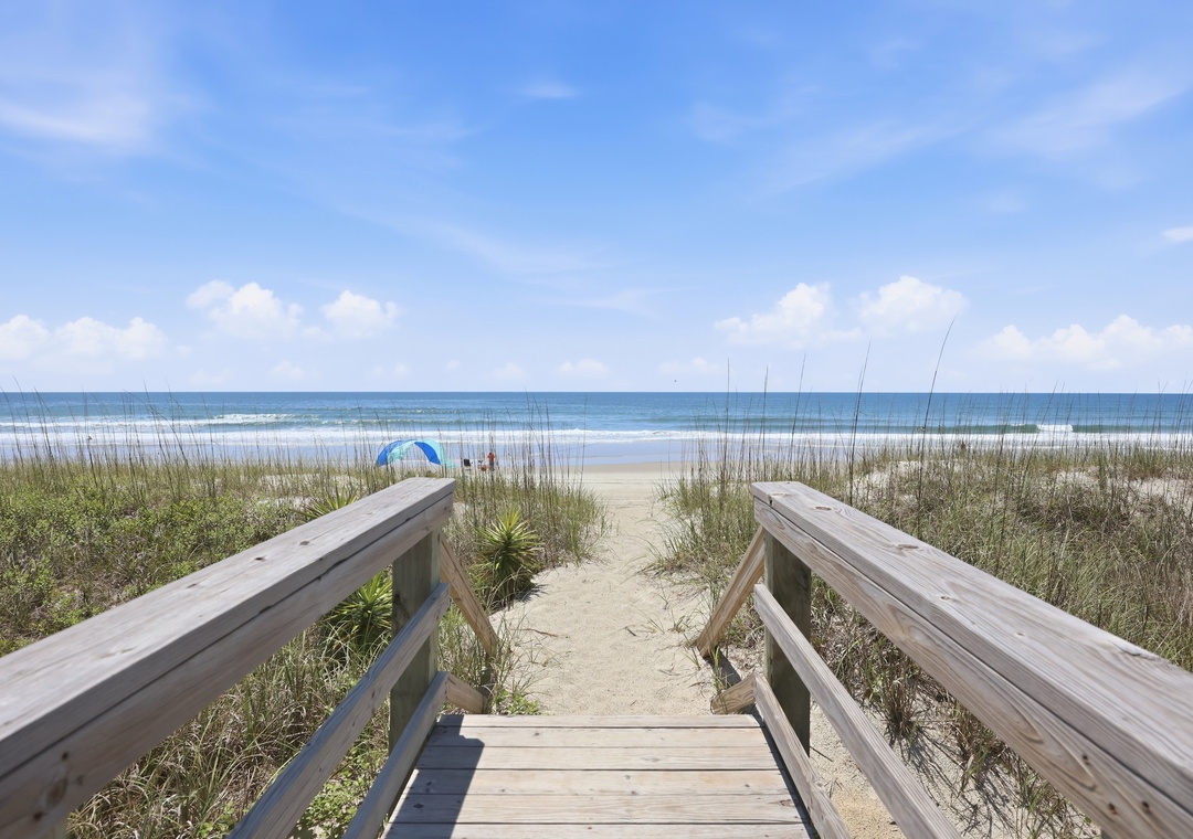 A private wooden boardwalk leads through gentle dunes, offering direct access to the wide sandy shore and sparkling ocean waves beyond.