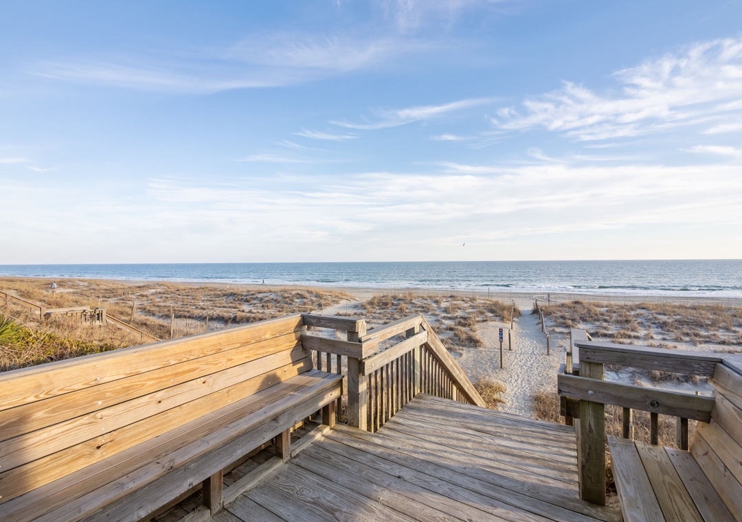 Wooden boardwalk leads through coastal dunes to pristine oceanfront beach under expansive sky.