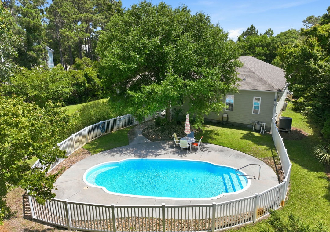 Aerial view showcasing a private backyard oasis with kidney-shaped pool, surrounded by lush greenery and mature trees. The property features a secure white fence, spacious lawn, and shaded patio seating.