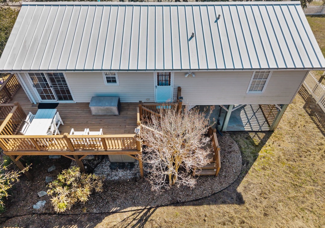 Charming beach house with metal roofing and wraparound deck, surrounded by natural landscaping and mature trees.