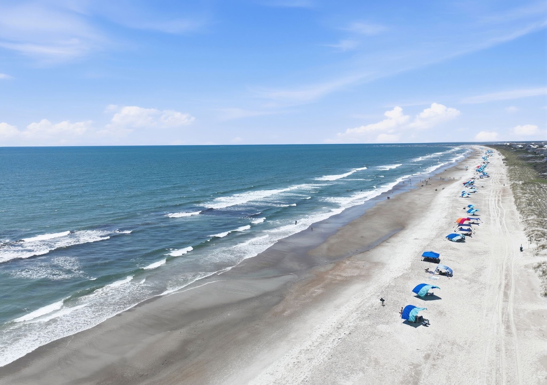 An aerial view captures a pristine stretch of sandy beach along the Atlantic coastline, with colorful umbrellas dotting the shore and gentle waves rolling in under a blue sky.