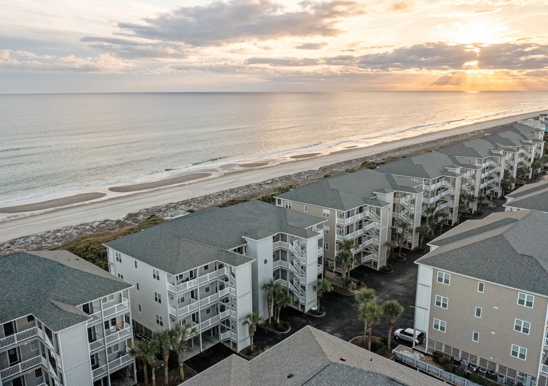 Beachfront resort buildings line pristine sandy shores under golden sunset skies, creating an idyllic coastal retreat setting.