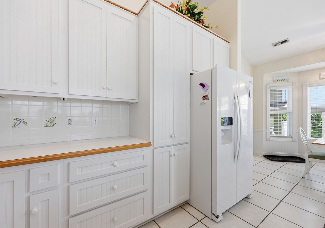 This bright, airy kitchen features classic white cabinetry, clean tile counters, and modern appliances ready for your culinary adventures. Natural light streams through windows overlooking peaceful greenery outside.