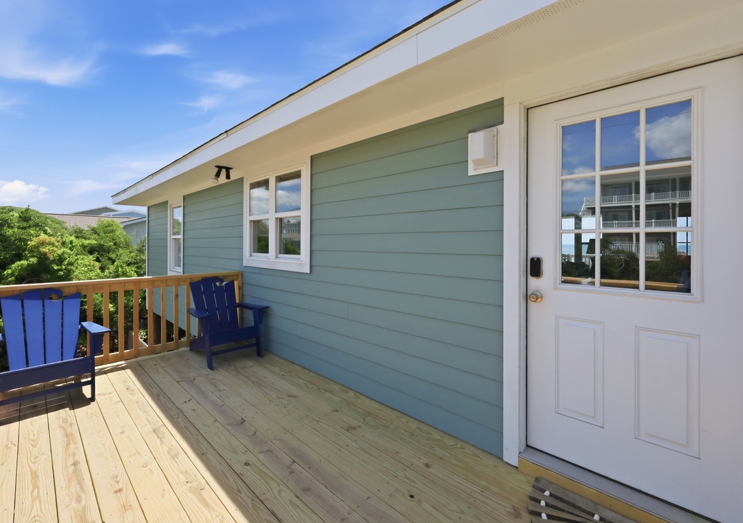A welcoming coastal-style deck with blue Adirondack chairs and glass-paneled entrance door, offering a bright outdoor retreat with natural surroundings.