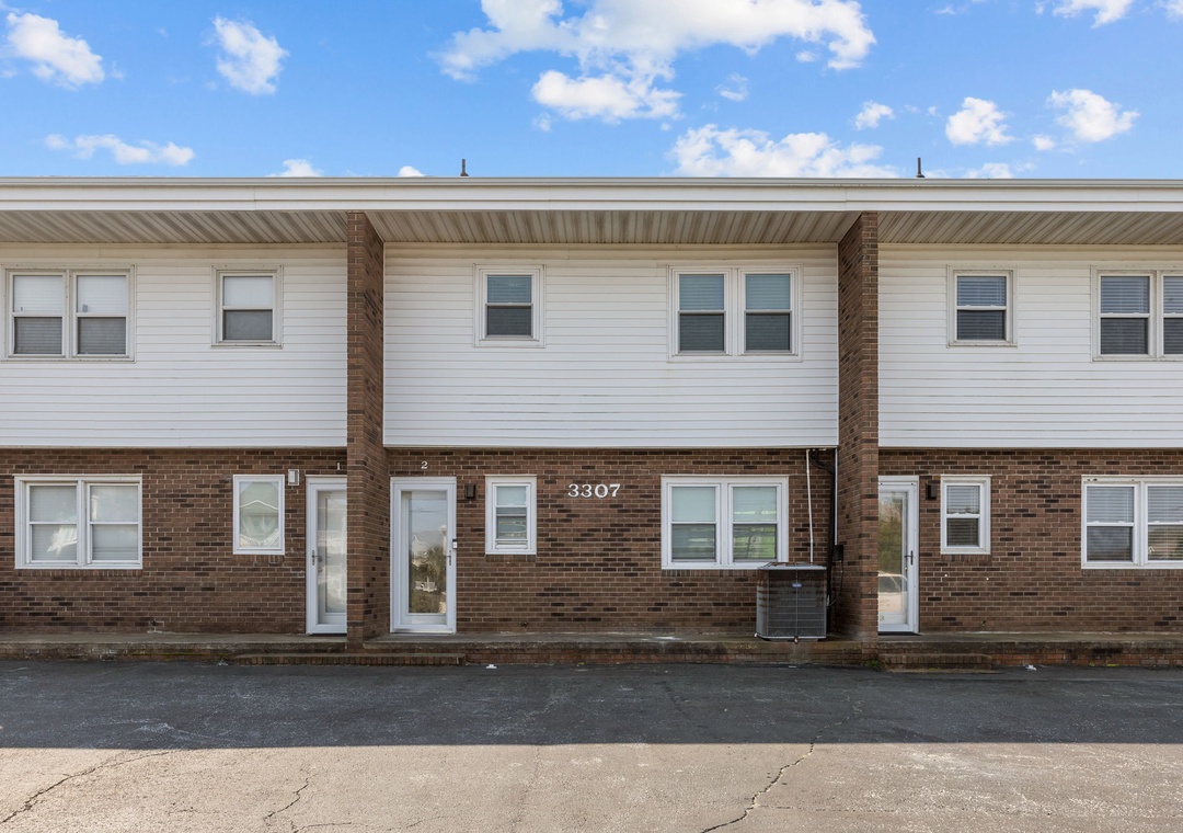 Multi-unit brick and vinyl siding building with individual entrances and parking area under bright blue skies.