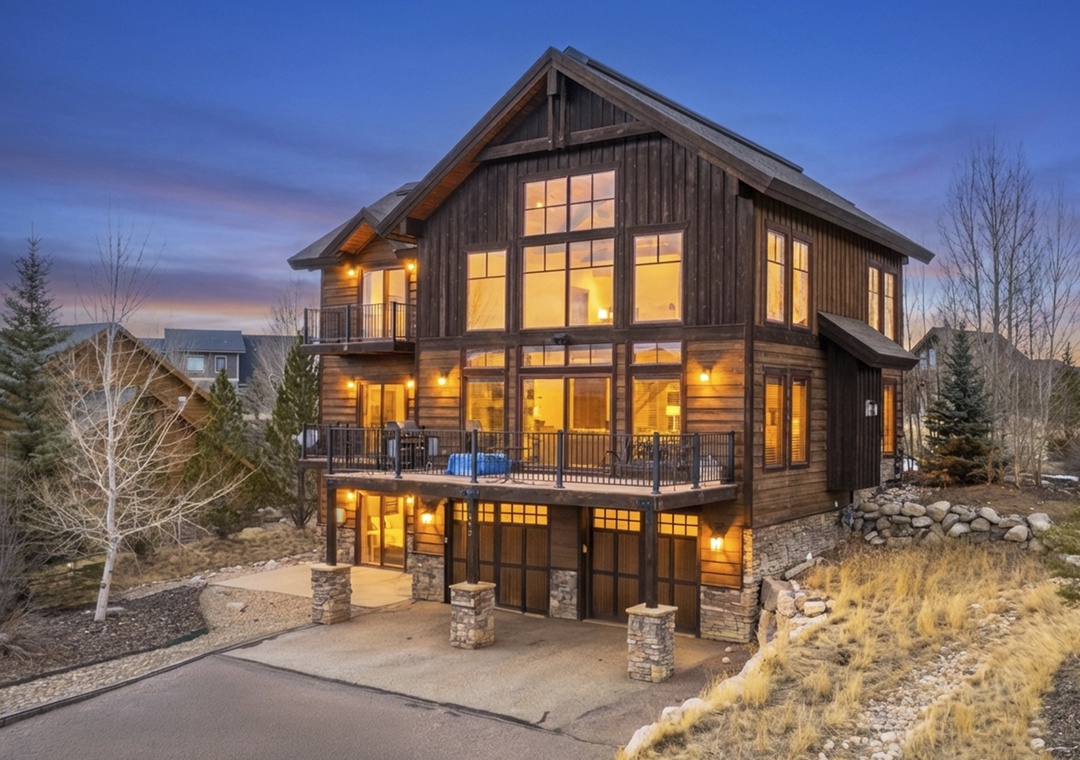 Stunning mountain lodge features warm wood exterior and expansive windows that glow invitingly at twilight in this peaceful neighborhood setting.