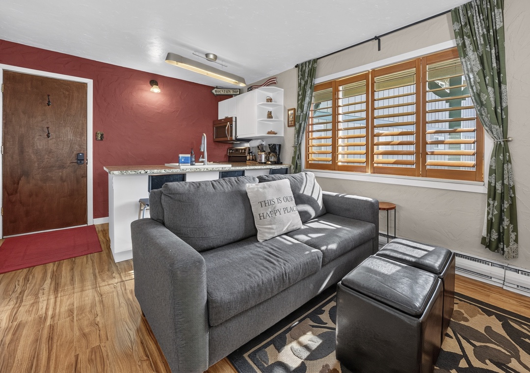 Sink into plush seating in your open living space, where warm red accent walls and natural light through plantation shutters create a cozy retreat. The kitchenette is steps away for easy meal prep.