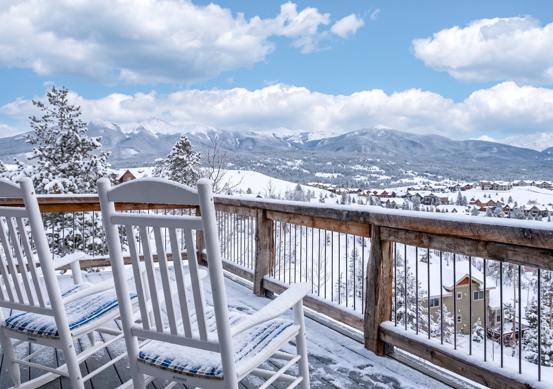 Two snow-covered white rocking chairs on a deck frame a majestic winter mountain landscape and village under a blue sky.