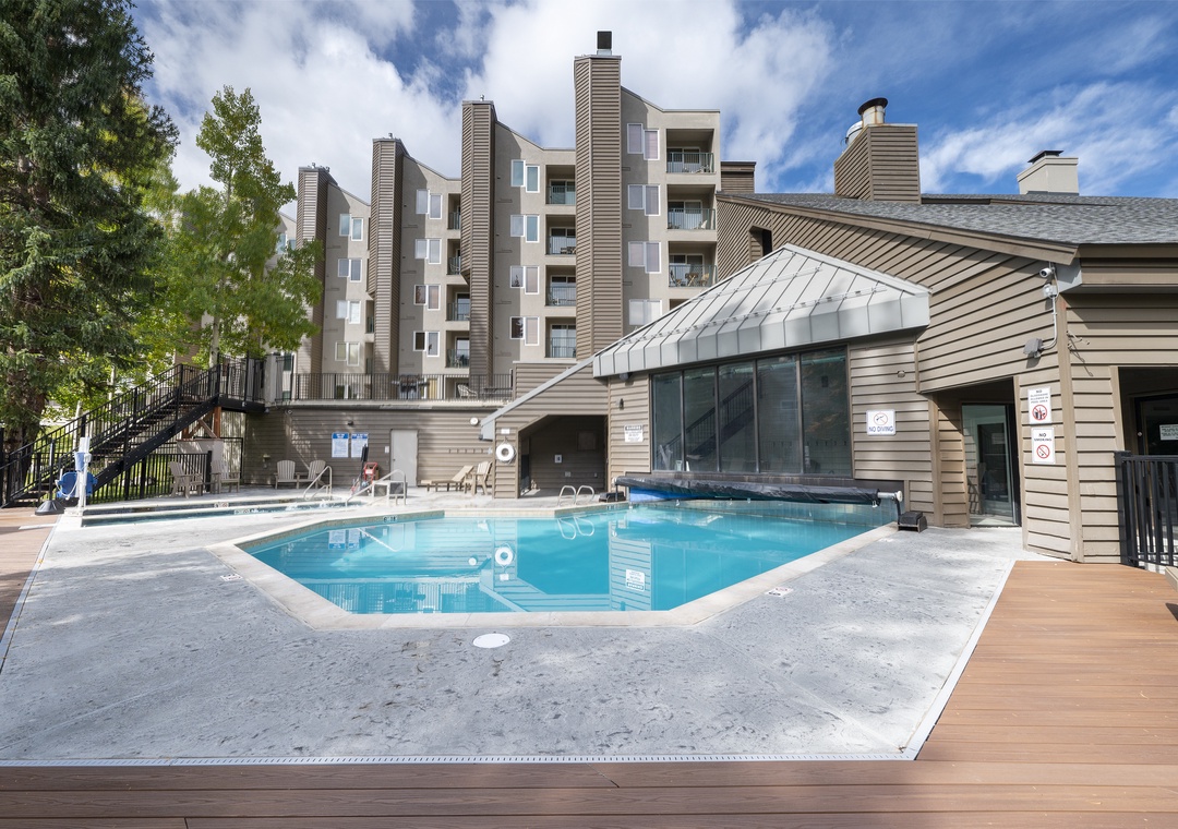 Outdoor pool area with mountain resort buildings and a covered hot tub pavilion under sunny skies.