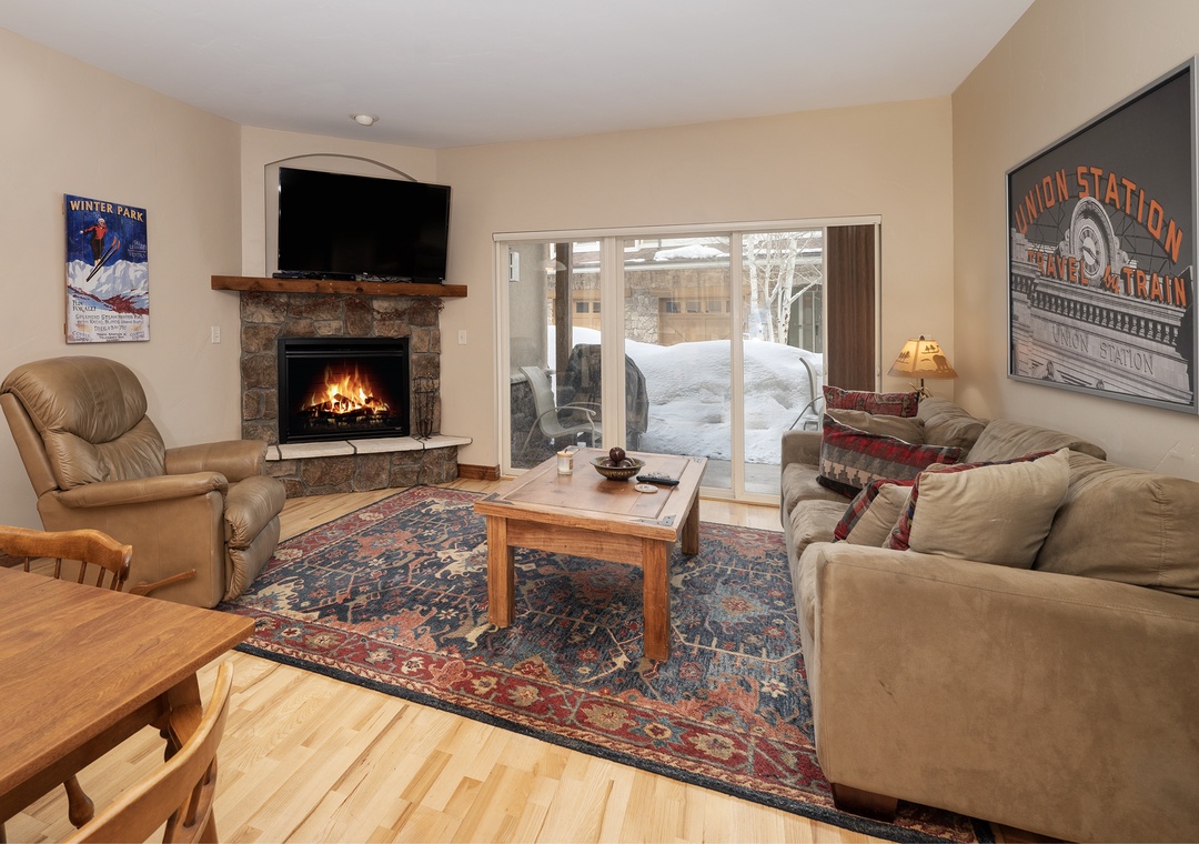 Cozy up in your warm living room with crackling fireplace, comfy seating, and snowy mountain views through sliding doors.