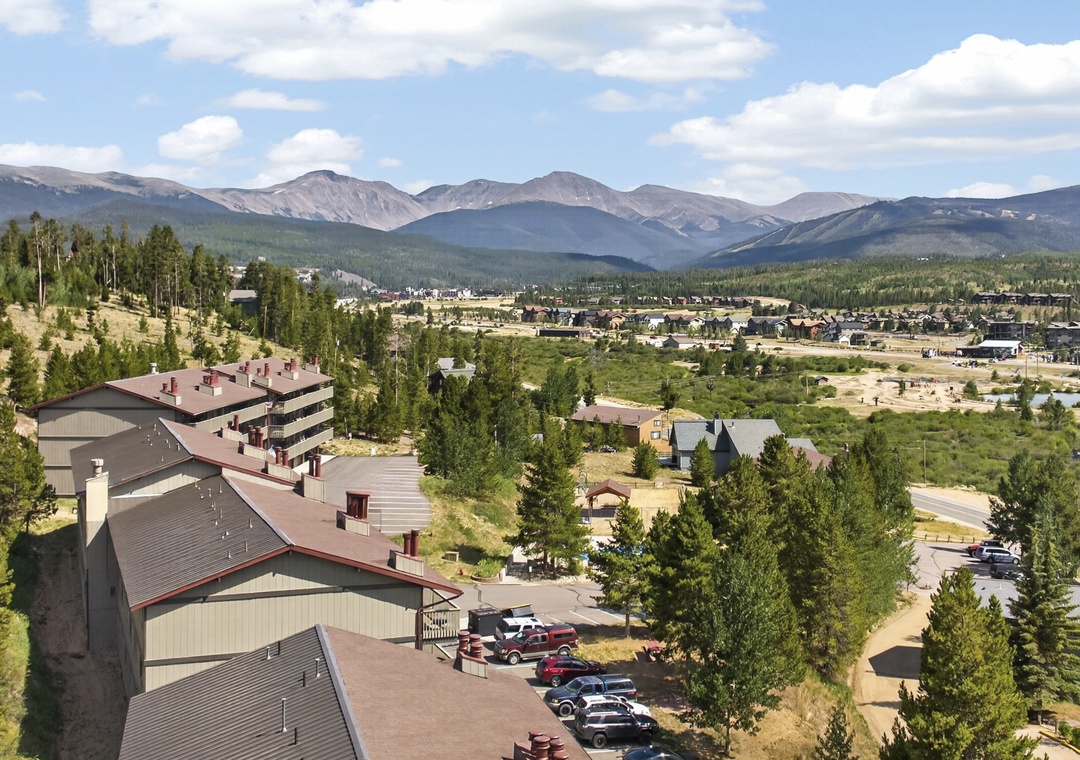 An elevated aerial view showcasing the property's mountain setting amid evergreen forests, with sweeping vistas of surrounding peaks and a nearby mountain town.