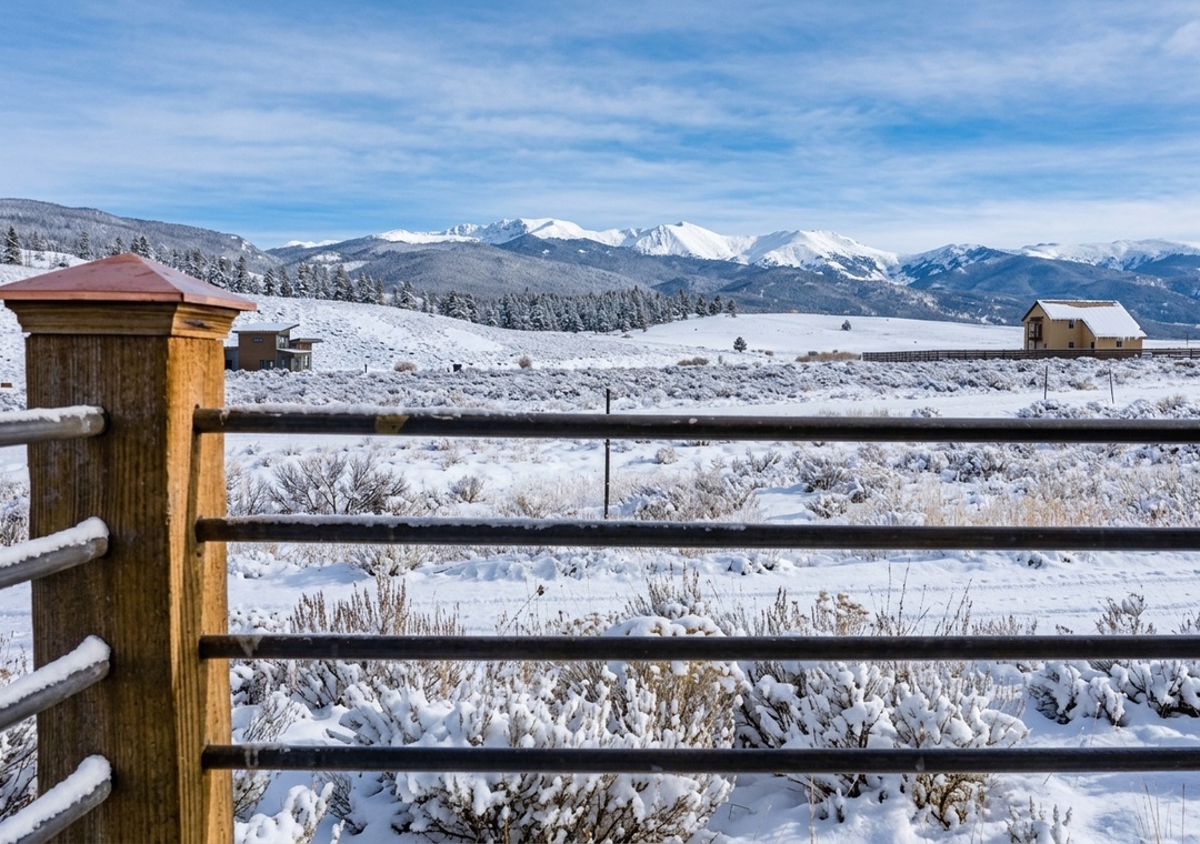 Dramatic mountain range rises beyond open fields and rustic ranch buildings, framing the property's peaceful countryside setting with sweeping natural beauty.