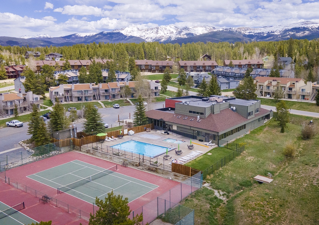 Aerial view of mountain resort community with recreational facilities including tennis court, swimming pool, and modern accommodations set against snow-capped peaks.
