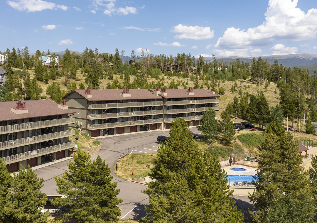 A mountain resort property nestled among evergreen forests, featuring multi-story buildings with balconies, outdoor parking, and a swimming pool, all set against a backdrop of distant mountain peaks and clear summer skies.