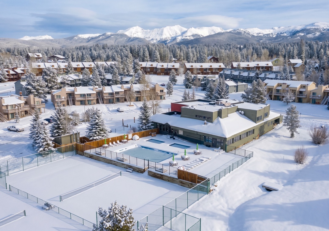 Aerial view of a mountain resort community blanketed in fresh snow, with snow-covered peaks and evergreen forests forming a stunning winter backdrop.