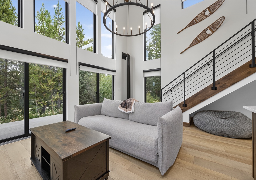 Soaring ceilings and floor-to-ceiling windows frame your peaceful forest view, while the cozy seating area invites you to unwind beneath a statement chandelier in this bright, airy living space.