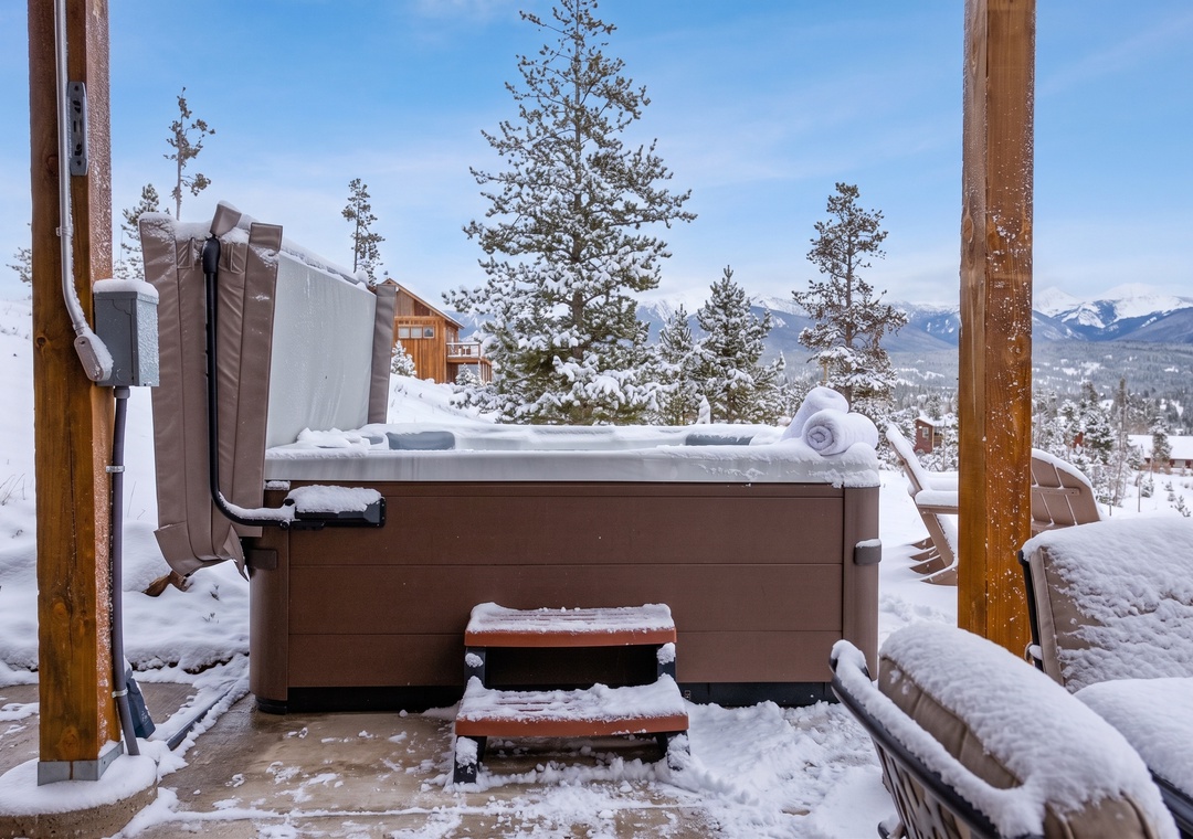 An outdoor hot tub sits open on a snow-covered patio, surrounded by snow-dusted pine trees and distant mountain views under a bright blue winter sky.
