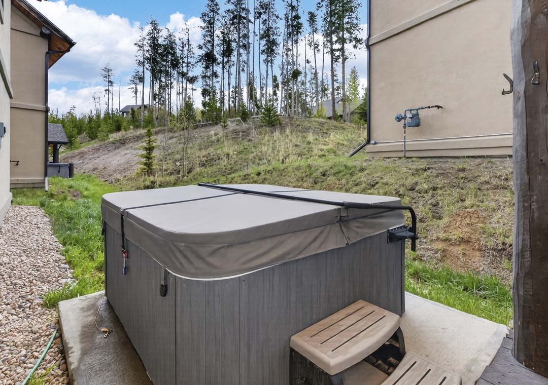A private hot tub sits on the back patio, offering mountain hillside views. The rental is nestled among tall evergreens in a peaceful natural setting.