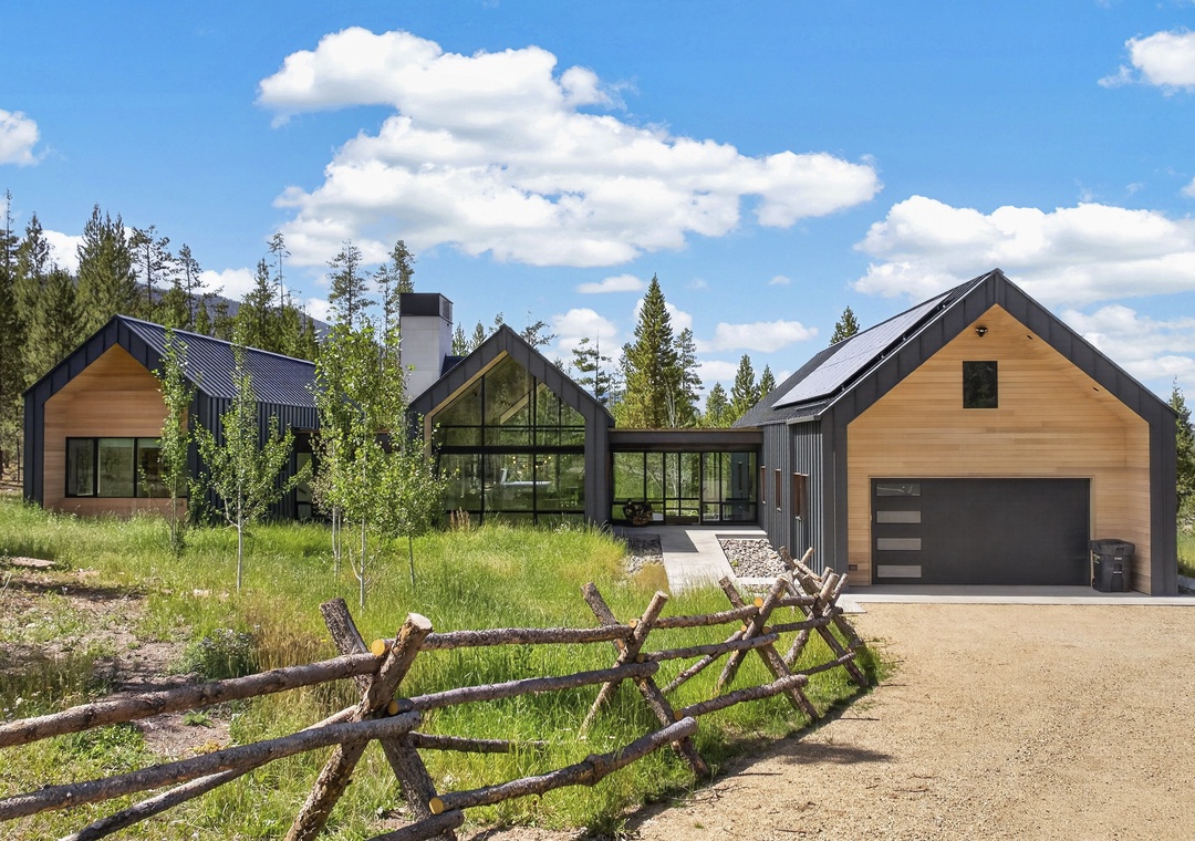 A striking modern mountain retreat set among tall pines and meadows, featuring a gravel driveway and rustic split-rail fence, offering a secluded escape in a natural landscape.