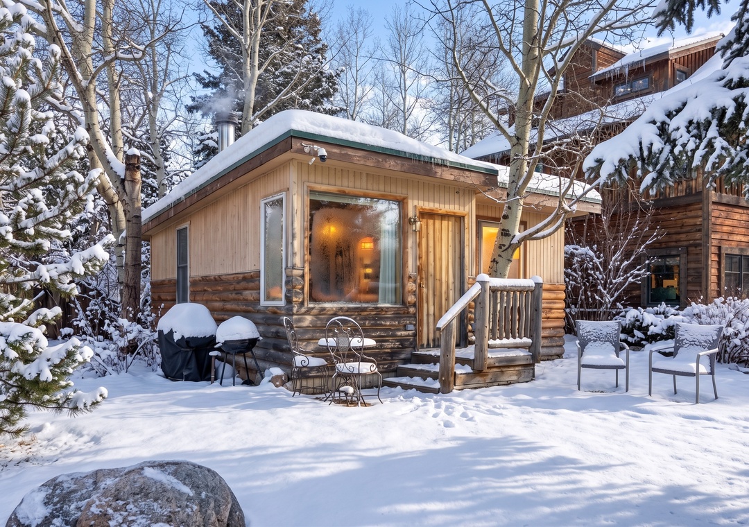 A cozy small wooden cabin nestled in a winter landscape, featuring a mix of vertical and log siding. Thick snow covers the roof, the surrounding trees, and the outdoor patio furniture, while warm light glows invitingly from the large front window.