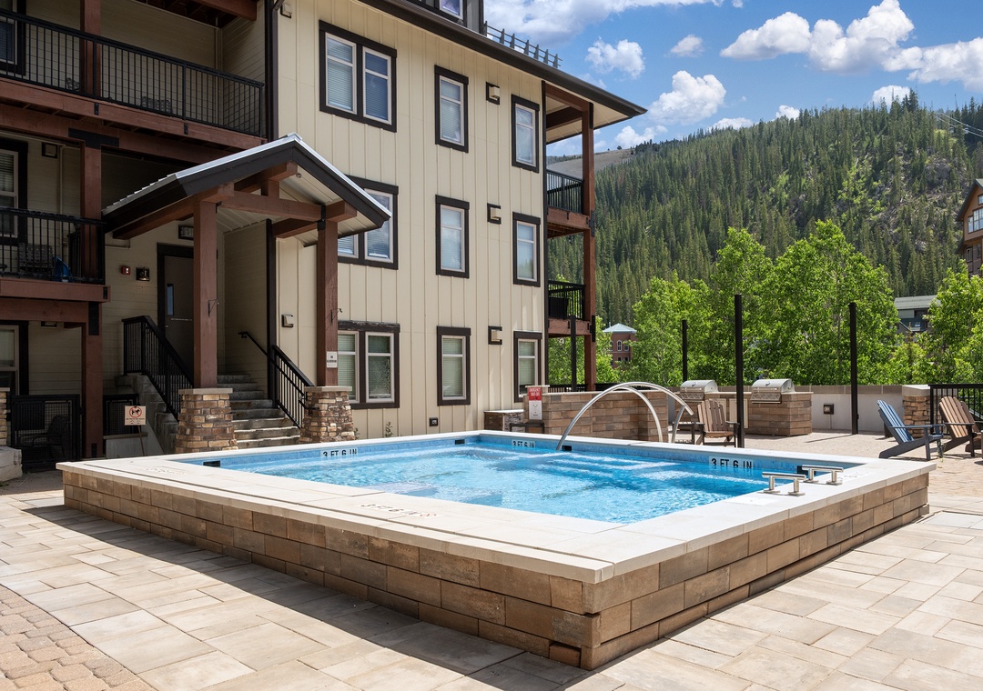 A multi-story property building surrounded by forested mountain views, featuring a raised hot tub and outdoor seating on a paved patio with modern exterior design.