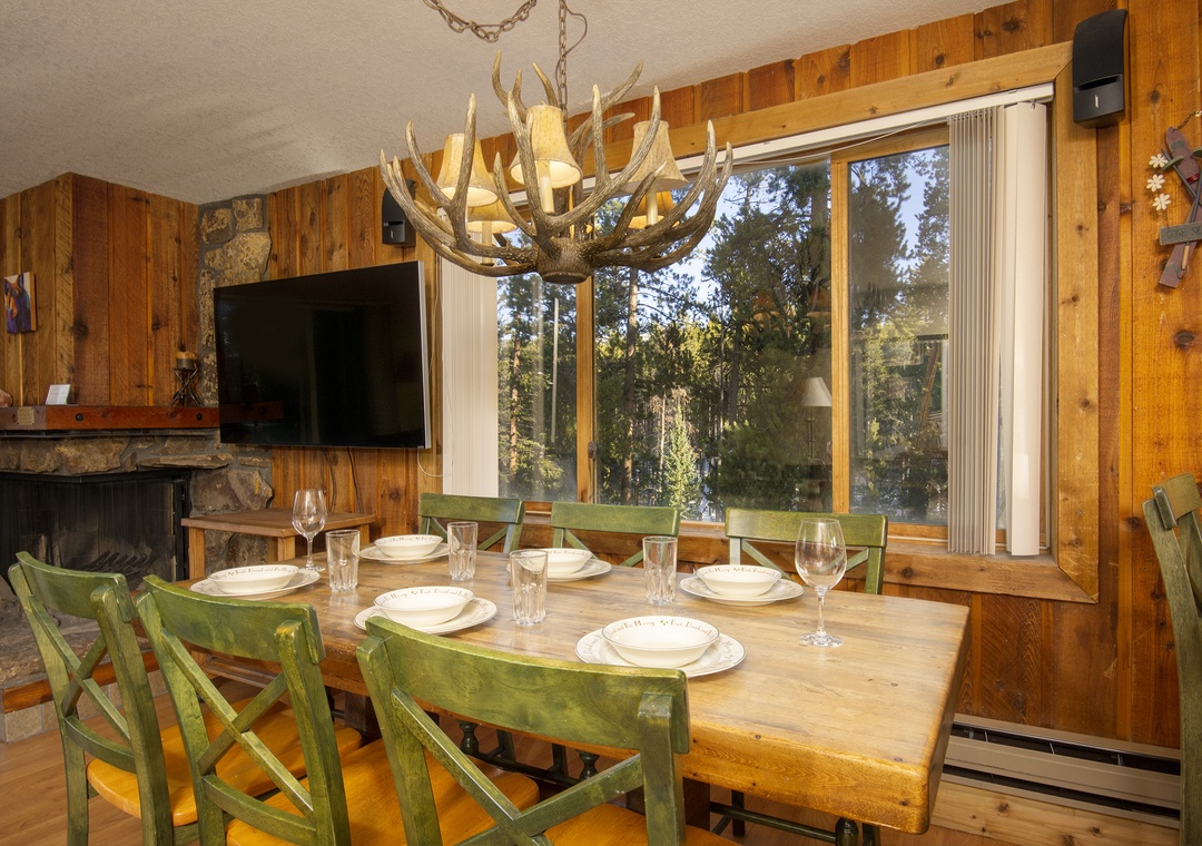 Gather around your rustic dining table beneath a stunning antler chandelier, where warm wood surrounds you and forest views create the perfect mountain retreat atmosphere.