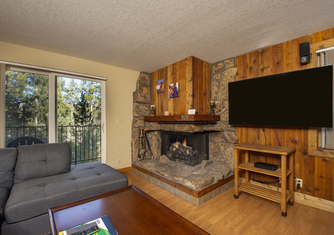 Cozy up by your stone fireplace while watching TV, with forest views through sliding doors creating the perfect mountain retreat atmosphere.