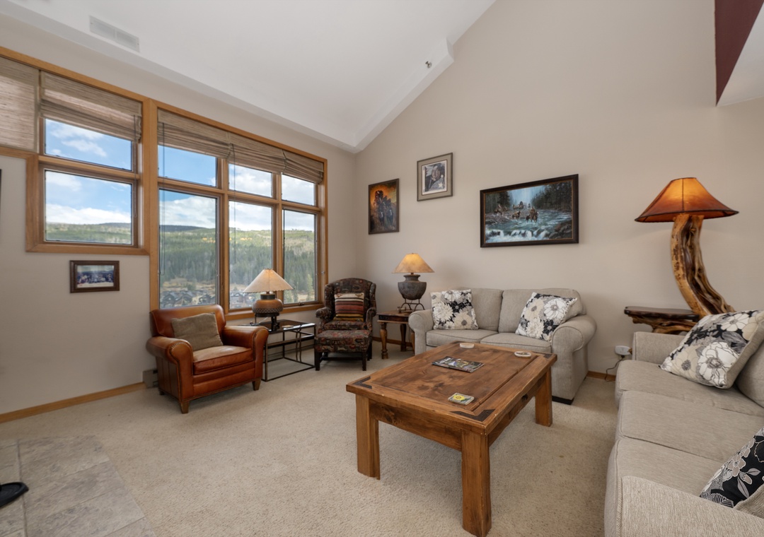 Relax in your mountain retreat's bright living space, where floor-to-ceiling windows frame stunning forest views and cozy furnishings invite you to unwind.