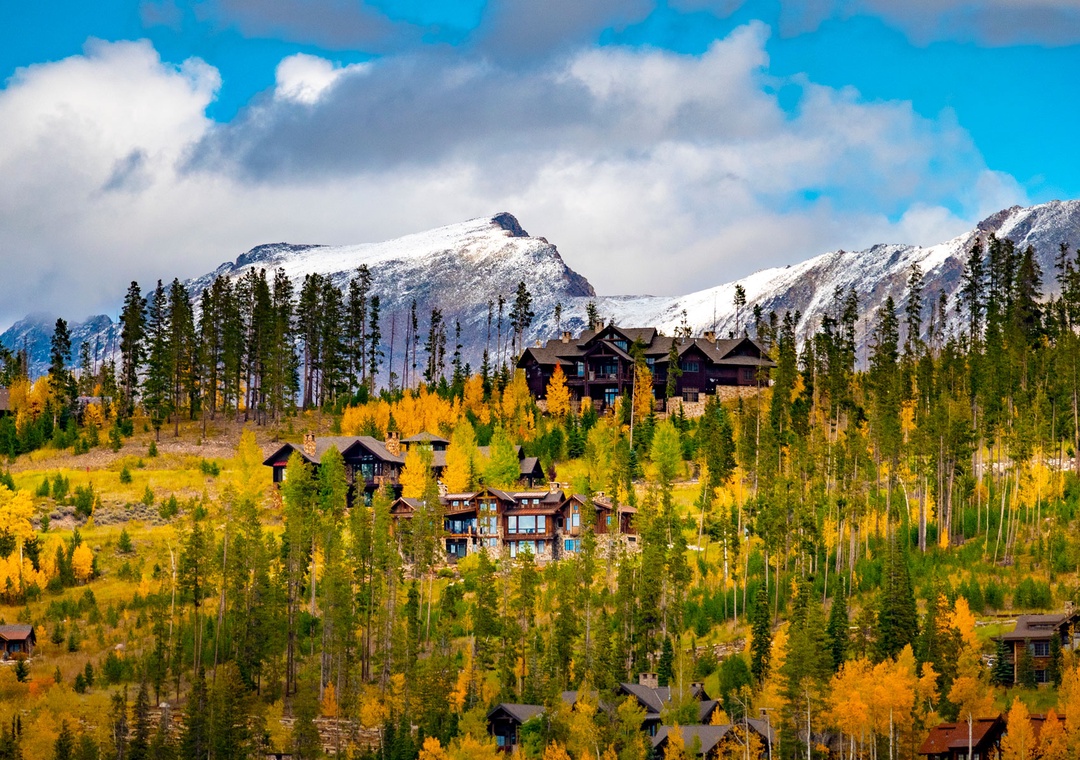 Mountain lodges nestled among golden aspens and evergreens with snow-capped peaks forming a dramatic backdrop.