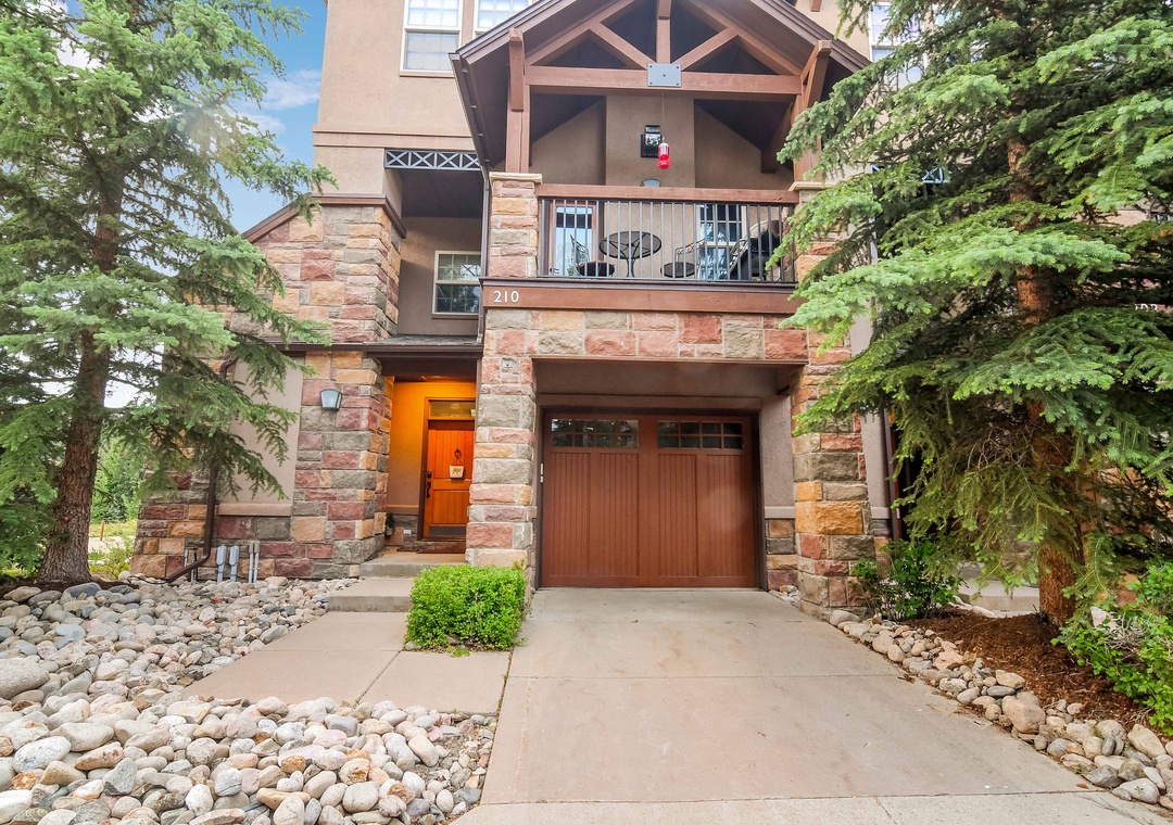 A charming mountain-style entrance framed by evergreens and natural stone, with private parking and a welcoming covered balcony above.