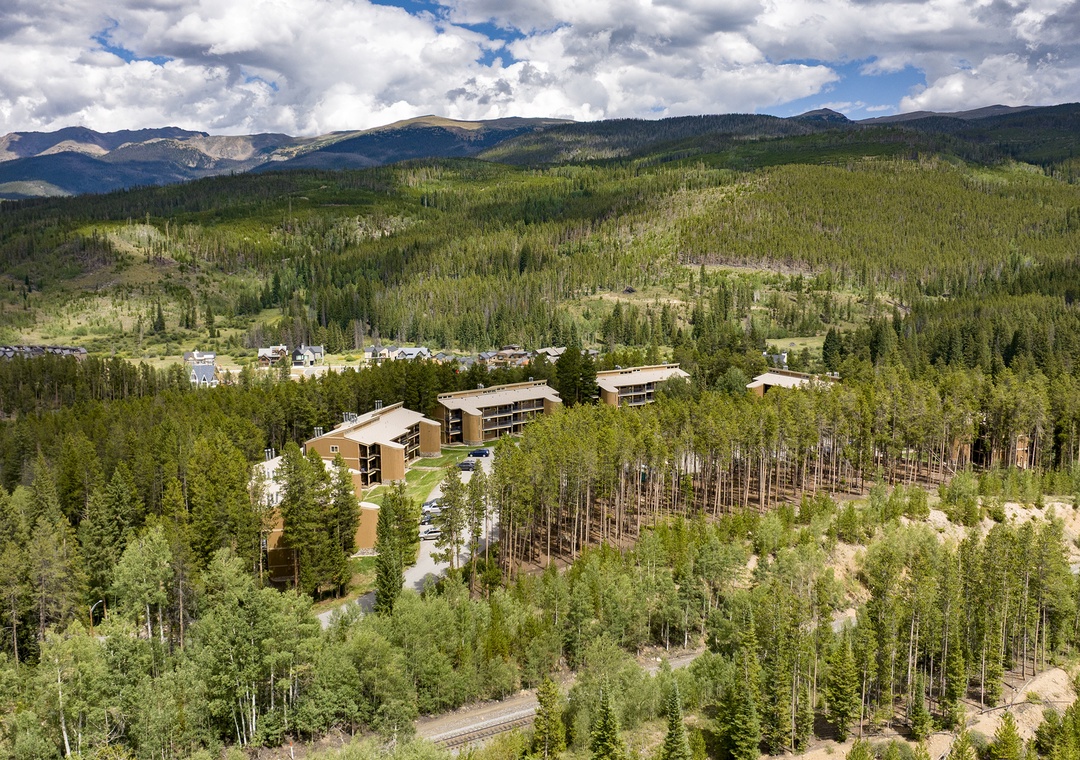 Aerial view of the property nestled among evergreens and aspen, with sweeping mountain ranges rising in the distance. The buildings rest within a vast natural landscape, offering direct access to the outdoors.