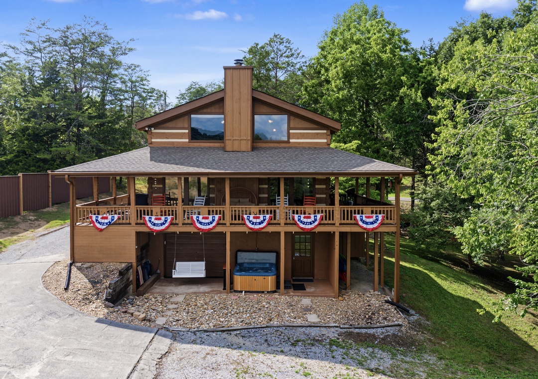 A welcoming mountain cabin features a wraparound deck adorned with patriotic bunting, a private hot tub, and ample parking, all nestled among towering trees in a peaceful wooded setting.