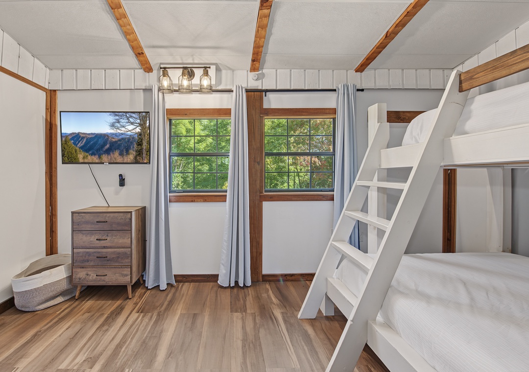 Cozy cabin-style retreat with warm wood beams, built-in bunk space perfect for families, and natural light from wood-framed windows. Polished wood flooring and rustic furnishings complete the welcoming atmosphere.