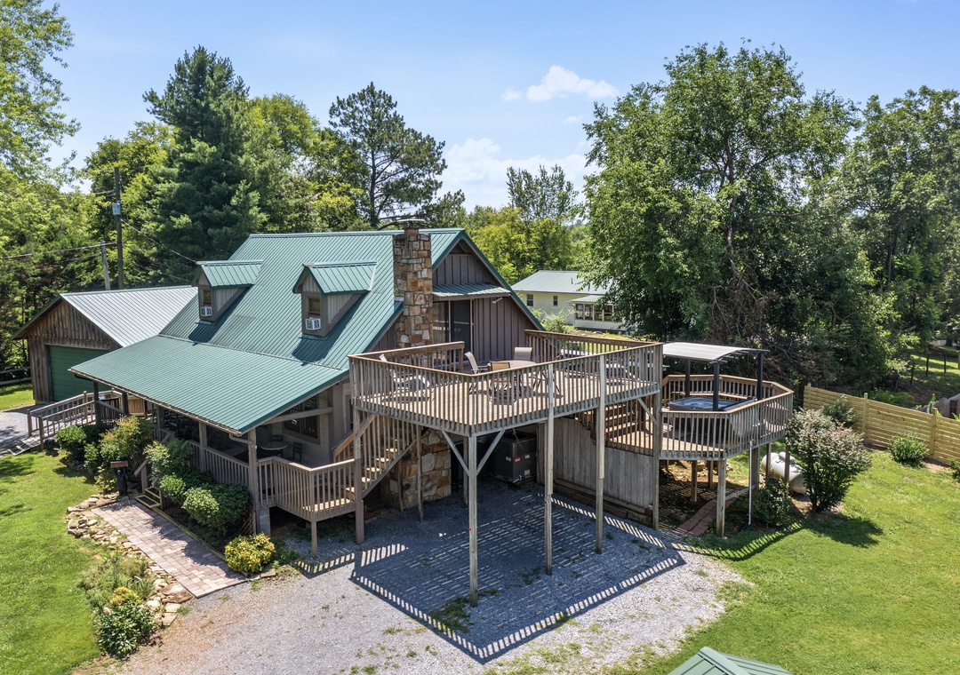 Aerial view of a spacious cabin retreat featuring distinctive green metal roofing, stone chimney, and expansive wraparound decks nestled within a lush, wooded setting with ample parking and outdoor areas.