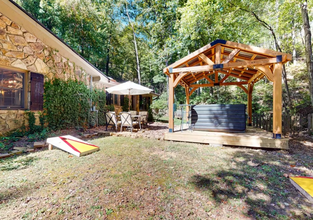 Backyard paradise with hot tub pavilion, cornhole game, and dining area surrounded by mature trees.