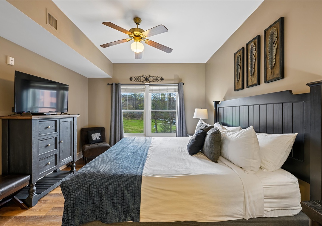 Relax in your comfortable bedroom with plush bedding, ceiling fan, and peaceful garden views through large windows.
