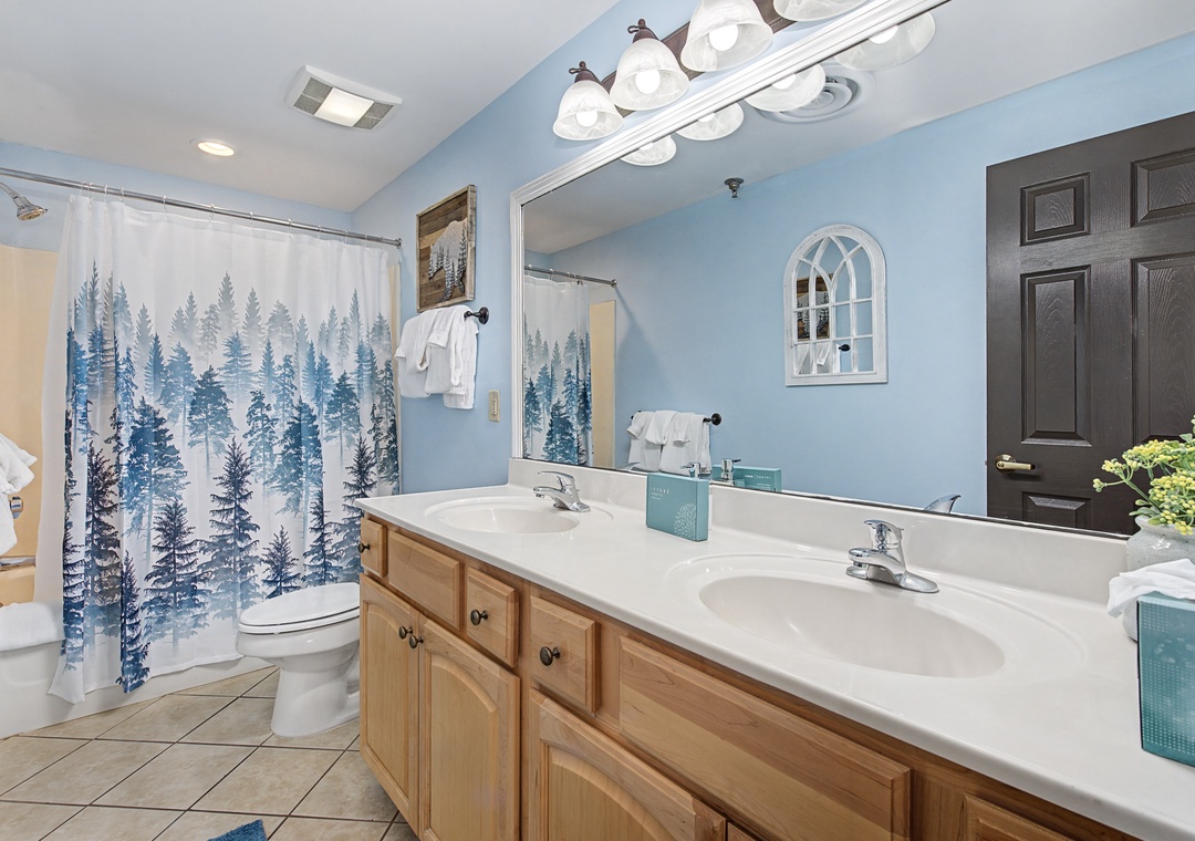 Your spacious bathroom features dual sinks, beautiful blue accents, and a relaxing forest-themed shower curtain to start your day refreshed.