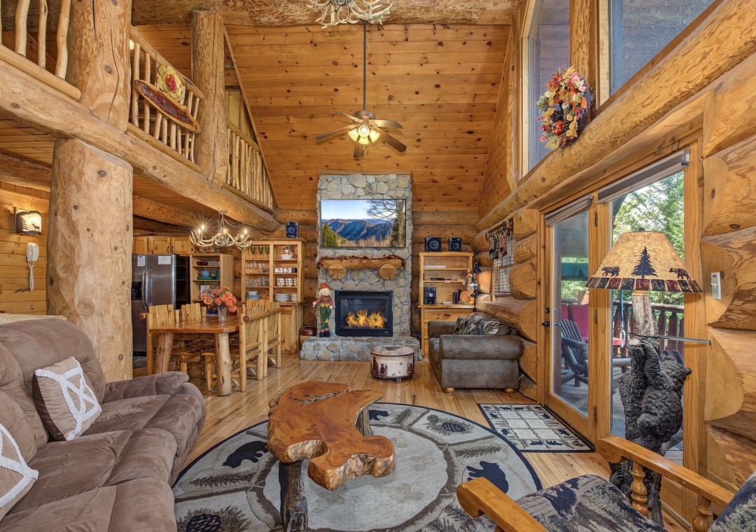 Cozy mountain retreat with rustic log beams, glowing stone fireplace, and large windows framing stunning views. Open layout flows seamlessly to the kitchen and outdoor deck.