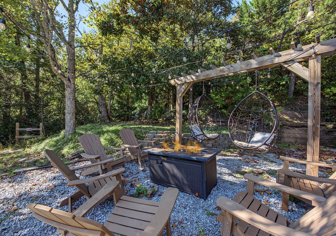 A tranquil outdoor retreat featuring a wooded setting with Adirondack chairs, hanging swing seats, and a firepit. The property is surrounded by lush greenery with string lights overhead.