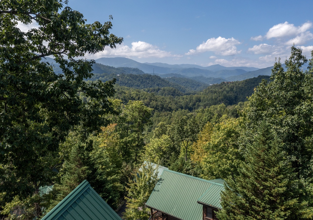 Stunning mountain vistas stretch across rolling forested hills beneath a bright blue sky dotted with white clouds.