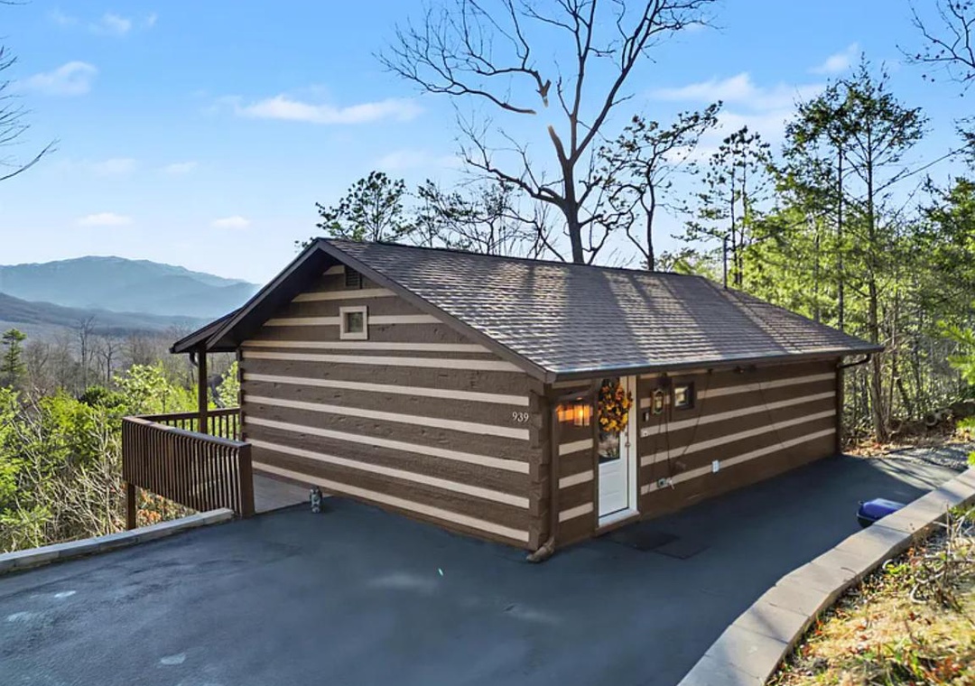 A rustic log cabin perched on a mountainside, offering panoramic views of layered ridges stretching into the distance. The property is nestled among tall trees and features a spacious deck and private parking area.