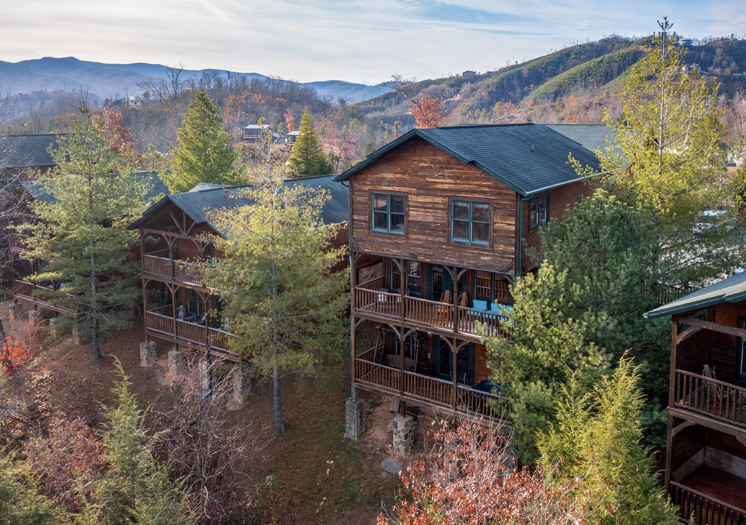 Aerial view of the property nestled in a forested mountain setting, showcasing the rustic wooden exterior and multi-level balconies surrounded by autumn foliage and scenic peaks.