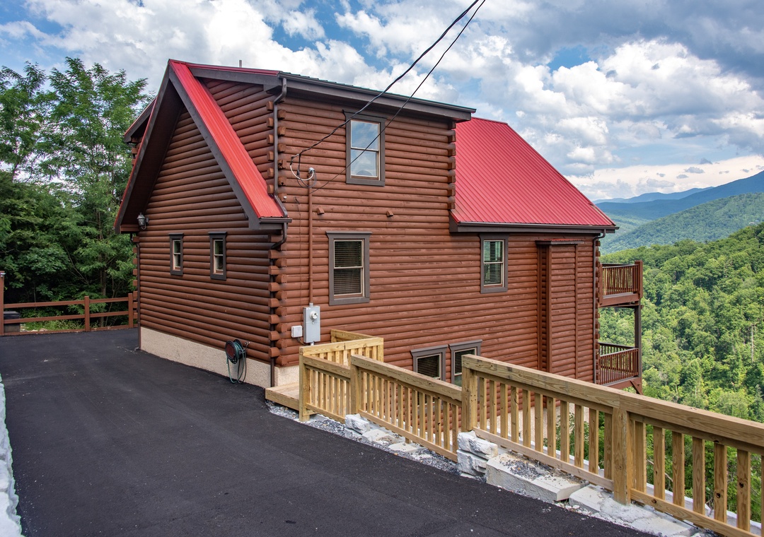 A charming log cabin with a striking red metal roof, nestled on a mountainside with sweeping forested valley views and convenient paved parking at the entrance.