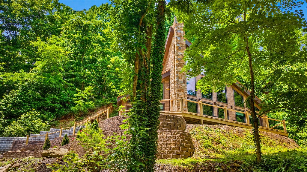 A charming cabin nestled in lush woods features a striking stone chimney and expansive windows framing the vibrant forest, creating a serene natural retreat.
