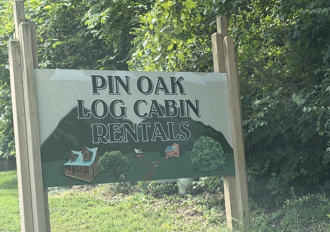 Pin Oak Log Cabin Rentals sign marks the entrance to the property, set against a backdrop of lush greenery and mature trees.