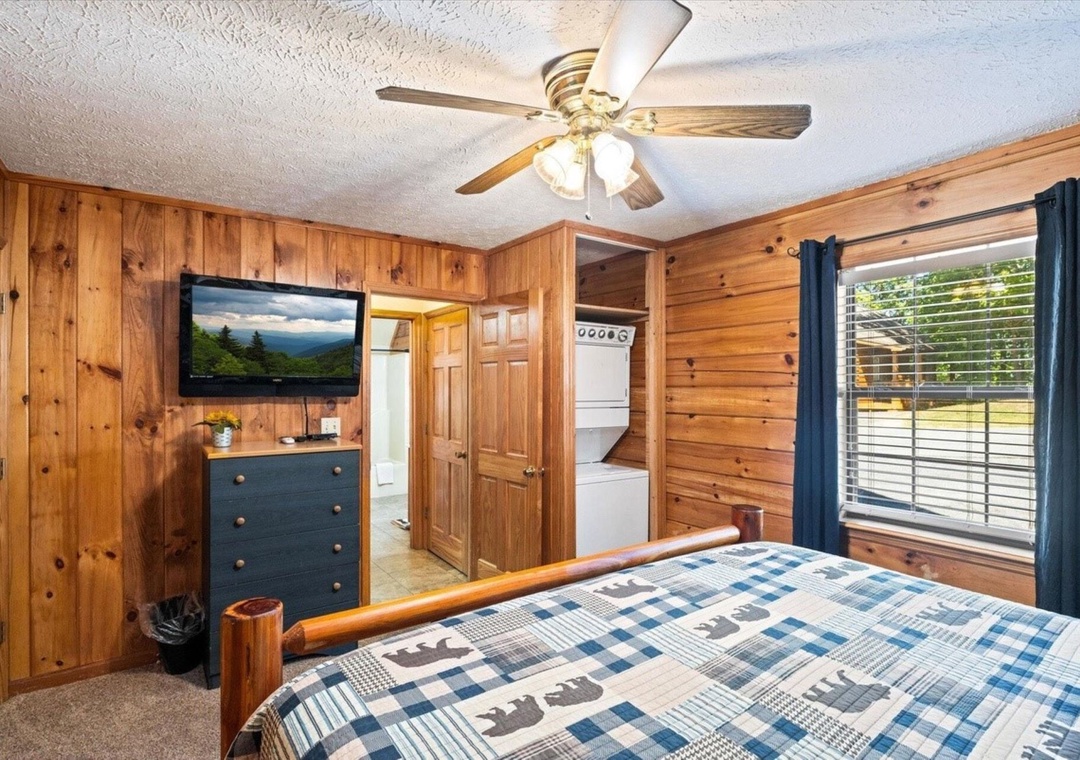 Your cozy cabin bedroom features rustic wood paneling, a comfy bed with mountain-themed bedding, and modern touches like TV and ceiling fan for perfect comfort.