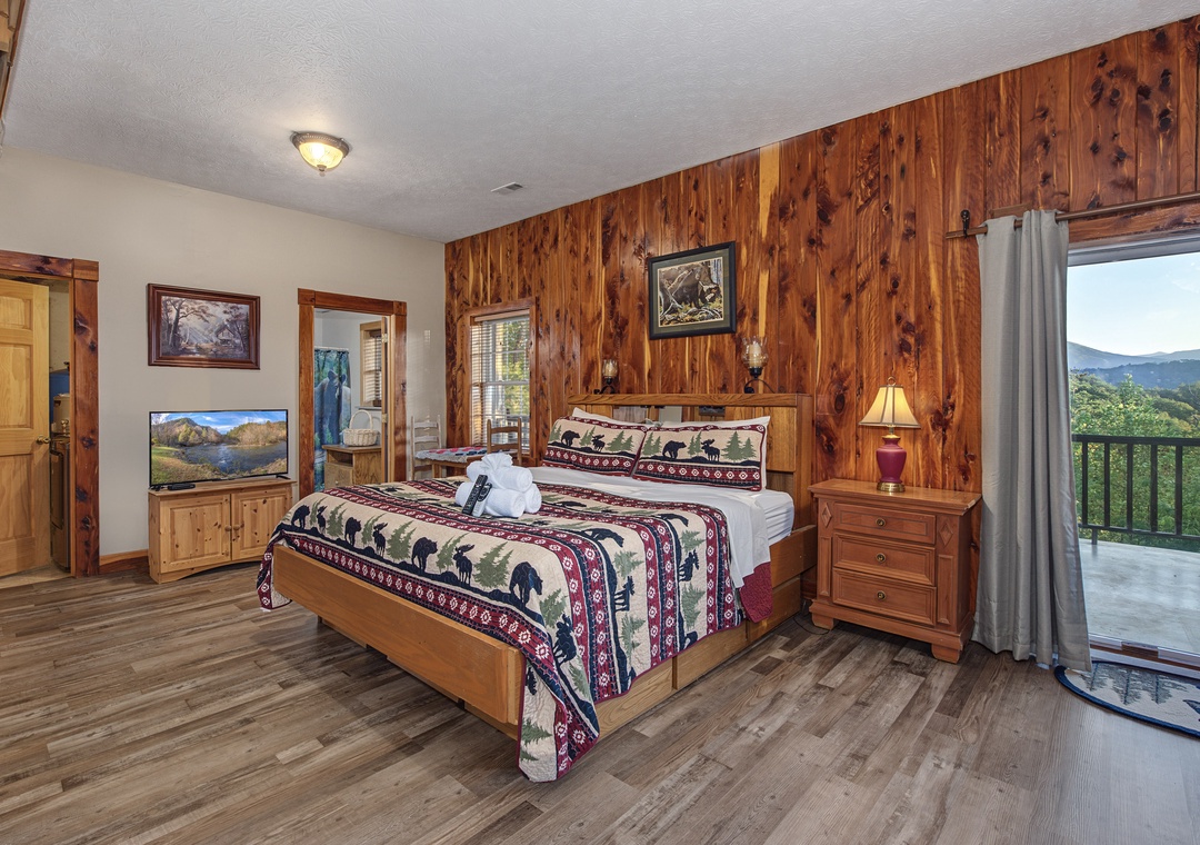 Step into your rustic retreat with warm wood-paneled walls and a cozy bed dressed in cabin-inspired linens. Through the glass door, your own private balcony frames stunning mountain views—perfect for your morning coffee or evening unwind.