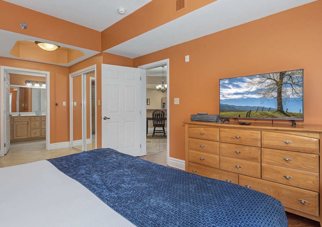 Unwind in your spacious master bedroom with warm orange walls, elegant coffered ceiling, and large TV for cozy evenings.