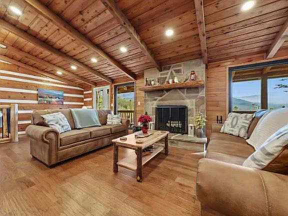 Relax in this inviting cabin living room with vaulted wood ceilings, a cozy stone fireplace, and plush seating bathed in warm natural light from large windows framing peaceful mountain views.