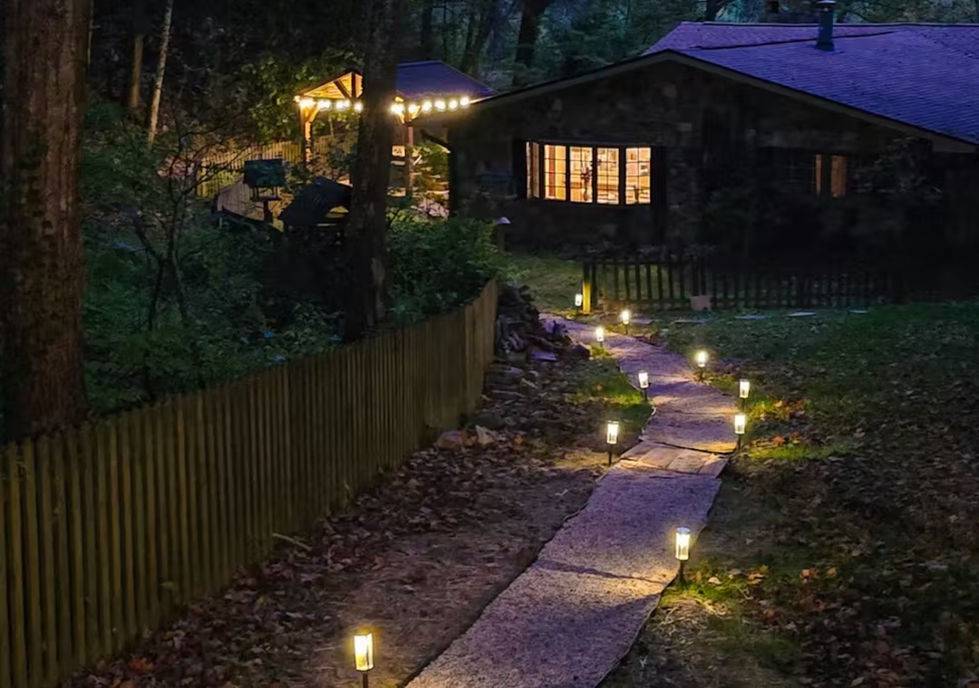 A charming woodland cabin glows warmly at dusk with illuminated pathways guiding your way through the peaceful forest setting.